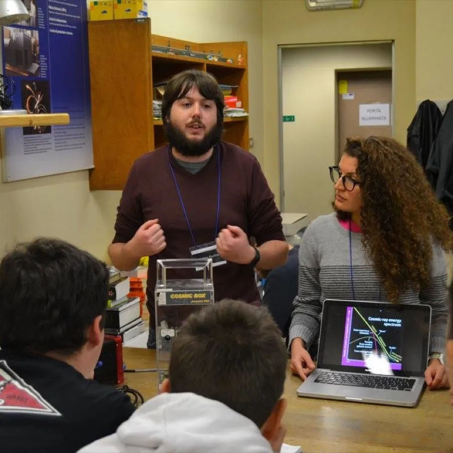 Matteo Nurisso explaining cosmic-ray physics to students during an outreach activity, with a cosmic-ray detector and a laptop on the table.